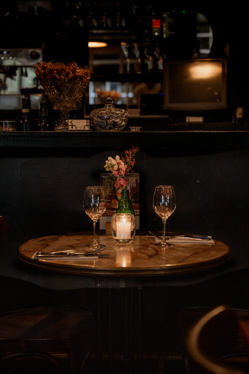Een rond tafeltje bij de bar, met kaarslicht, bloemen en twee wijnglazen